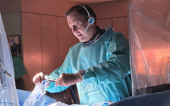 Arzt während einer elektrophysiologischen Behandlung im Herzkatheterlabor. In steriler Schutzkleidung arbeitet er an einem Kathetersystem, das über die Blutgefäße zum Herzen geführt wird. Moderne Medizintechnik und abgeschirmte Gerätebereiche sind im Hintergrund sichtbar. Das Bild veranschaulicht einen minimalinvasiven Eingriff zur Behandlung von Herzrhythmusstörungen.