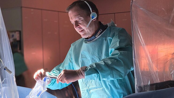 Arzt während einer elektrophysiologischen Behandlung im Herzkatheterlabor. In steriler Schutzkleidung arbeitet er an einem Kathetersystem, das über die Blutgefäße zum Herzen geführt wird. Moderne Medizintechnik und abgeschirmte Gerätebereiche sind im Hintergrund sichtbar.
